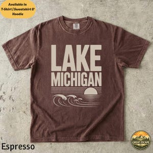 K&ouml;nnte beinhalten: Ein kurz&auml;rmeliges T-Shirt mit Rundhalsausschnitt in verwaschenem Espresso-Braun. Auf der Vorderseite befindet sich der Schriftzug LAKE MICHIGAN in gro&szlig;en beigen Gro&szlig;buchstaben &uuml;ber einer Grafik einer untergehenden Sonne und stilisierten Wellen.