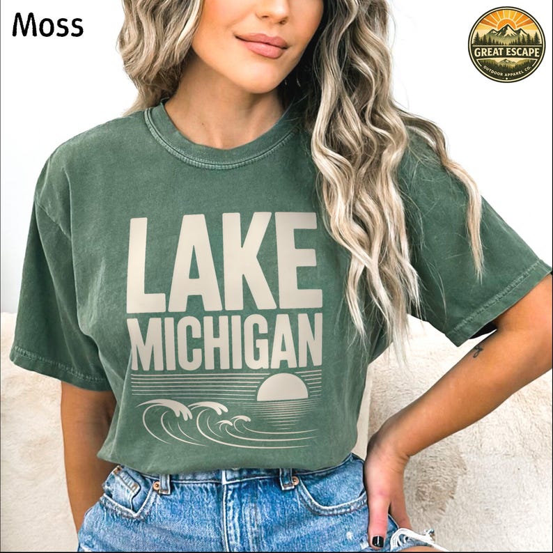 K&ouml;nnte beinhalten: Ein moosgr&uuml;nes Kurzarm-T-Shirt mit dem Text LAKE MICHIGAN in hellbeiger Schrift. Unter dem Text befindet sich eine Grafik einer untergehenden Sonne &uuml;ber stilisierten Wellen. Das T-Shirt hat einen Rundhalsausschnitt und eine Vintage-Waschung.