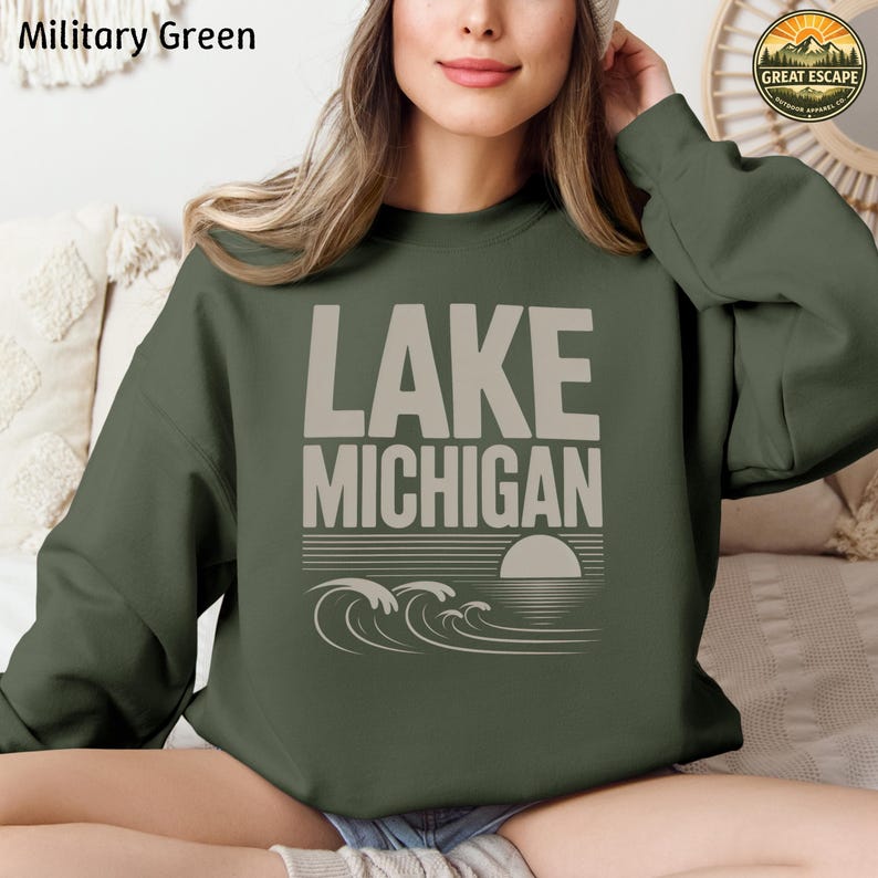 K&ouml;nnte beinhalten: Ein milit&auml;rgr&uuml;nes Langarm-Sweatshirt mit dem Text LAKE MICHIGAN in gro&szlig;en beigen Buchstaben &uuml;ber einer Grafik aus stilisierten Wellen und einer untergehenden Sonne. Das Design ist mittig auf der Brust des Kleidungsst&uuml;cks platziert.