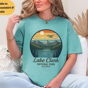以下が含まれることがあります： 山岳湖の風景を描いた円形グラフィックと「Lake Clark National Park Alaska」の文字が入った、シーフォームグリーンのTシャツ。 Tシャツ、スウェットシャツ、パーカーのスタイルがあります。