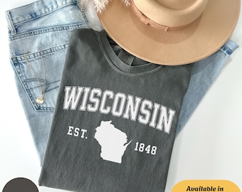 May include: A flat lay featuring a gray t-shirt with "WISCONSIN" printed in white, along with the state outline, "EST. 1848". The shirt rests on light blue jeans, with a tan hat and a sprig of dried flowers. Text on the image says "Available in T-Shirt / Sweatshirt & Hoodie".