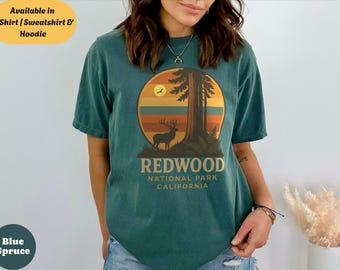 May include: Teal t-shirt featuring a Redwood National Park design. The graphic includes a deer, a large tree, and the text "REDWOOD NATIONAL PARK CALIFORNIA" in a circular design with a sunset background. Available in T-Shirt, Sweatshirt & Hoodie.