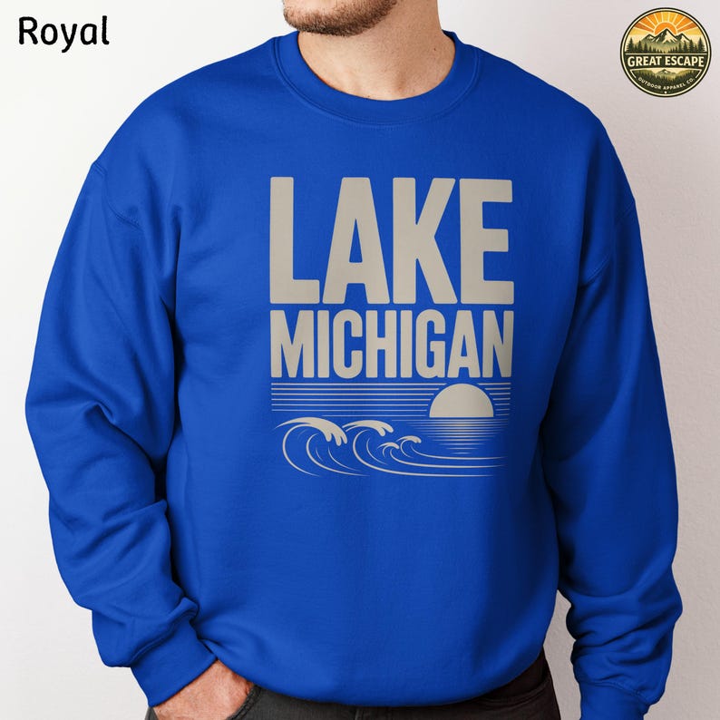 K&ouml;nnte beinhalten: Ein k&ouml;nigsblaues Sweatshirt mit Rundhalsausschnitt und dem Text LAKE MICHIGAN in gro&szlig;en beigen Buchstaben. Unter dem Text befindet sich eine Grafik einer untergehenden Sonne und stilisierter Wellen in Beige. Ein rundes Logo oben rechts zeigt GREAT ESCAPE.