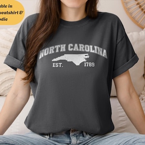 May include: Dark gray t-shirt featuring the text "NORTH CAROLINA" above a white outline of the state, with "EST. 1789" below. The shirt is available in T-shirt, sweatshirt, and hoodie styles.