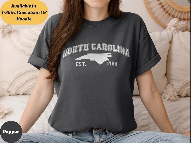 May include: Dark gray t-shirt featuring the text "NORTH CAROLINA" above a white outline of the state, with "EST. 1789" below. The shirt is available in T-shirt, sweatshirt, and hoodie styles.