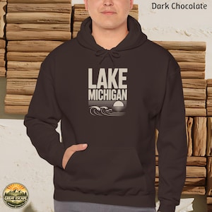 K&ouml;nnte beinhalten: Ein dunkel schokoladenbrauner Kapuzenpullover mit Kapuze, passenden Kordelz&uuml;gen und einer K&auml;ngurutasche. Auf der Brust befindet sich ein Aufdruck mit dem Text LAKE MICHIGAN &uuml;ber einem stilisierten Sonnen- und Wellenmotiv in Cremewei&szlig;.