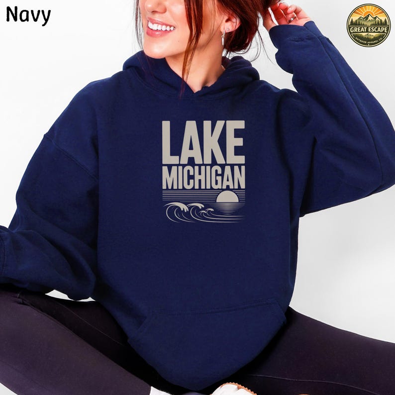 K&ouml;nnte beinhalten: Ein marineblauer Kapuzenpullover mit einem Grafikdruck auf der Brust, der den beigen Schriftzug LAKE MICHIGAN sowie eine stilisierte Darstellung von Wellen und einer untergehenden Sonne &uuml;ber dem Wasser zeigt, inklusive einer K&auml;ngurutasche.