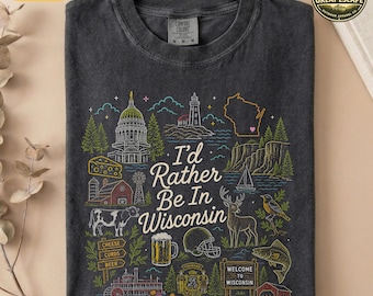 May include: A charcoal gray t-shirt featuring a multi-colored graphic print with the text "I'd Rather Be In Wisconsin." The design includes illustrations of a capitol building, a lighthouse, a cow, a deer, a beer mug, and a football helmet.