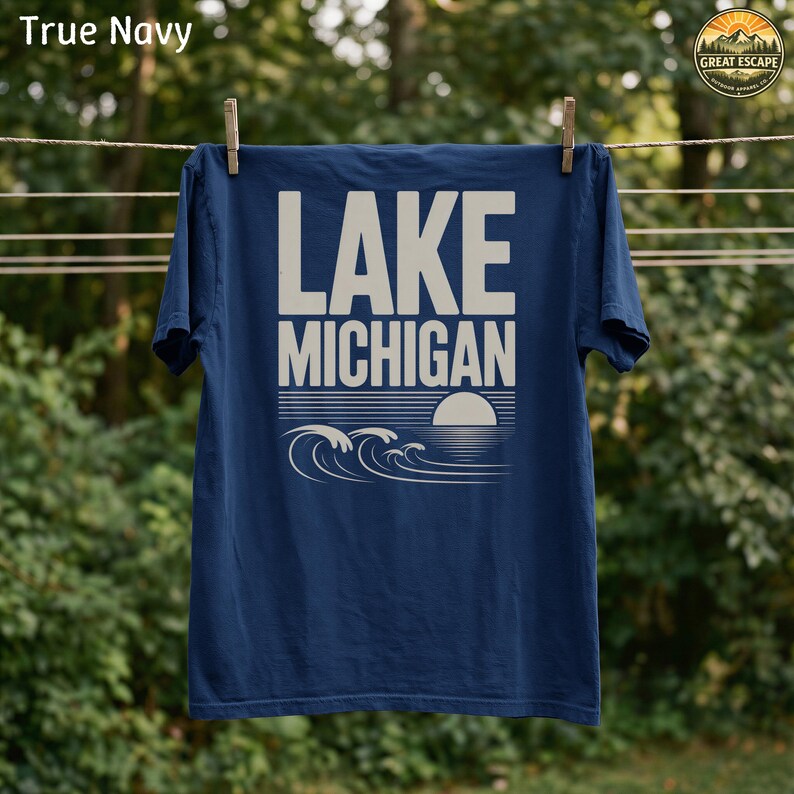 K&ouml;nnte beinhalten: Ein marineblaues T-Shirt mit kurzen &Auml;rmeln und dem Schriftzug LAKE MICHIGAN in gro&szlig;en cremefarbenen Buchstaben. Unter dem Text befindet sich eine grafische Illustration von stilisierten Wellen und einer untergehenden Sonne in Creme.