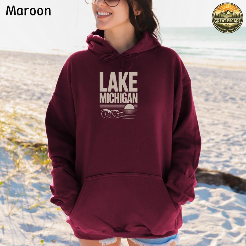 K&ouml;nnte beinhalten: Ein kastanienbrauner Kapuzenpullover mit dem Text LAKE MICHIGAN in hellbeiger Schrift. Unter dem Text befindet sich eine grafische Darstellung stilisierter Wellen und einer untergehenden Sonne am Horizont. Der Hoodie hat eine K&auml;ngurutasche.