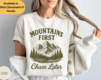 Può includere: T-shirt color avorio con la scritta "Mountains First Chaos Later" in verde oliva. Il design presenta una catena montuosa e alberi. Disponibile in T-Shirt, Felpa e Felpa con cappuccio.