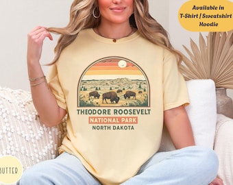 May include: Butter-colored t-shirt featuring a graphic of Theodore Roosevelt National Park. The design includes bison, a sunset, and the park's name. Text on the shirt reads "Available in T-Shirt / Sweatshirt & Hoodie."