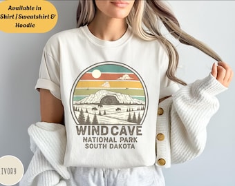May include: Ivory-colored t-shirt featuring a circular graphic of Wind Cave National Park in South Dakota. The design includes a mountain scene with bison, trees, and a sunset. Text reads "WIND CAVE NATIONAL PARK SOUTH DAKOTA". Available in T-Shirt, Sweatshirt & Hoodie.