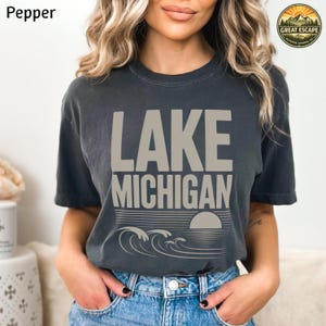 K&ouml;nnte beinhalten: Ein anthrazitfarbenes Kurzarm-T-Shirt mit dem Text LAKE MICHIGAN in gro&szlig;en beigen Buchstaben. Unter dem Text befindet sich eine Grafik einer untergehenden Sonne &uuml;ber stilisierten Meereswellen. Das T-Shirt hat einen Rundhalsausschnitt.