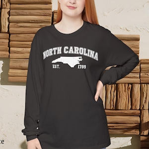 May include: Graphite long-sleeve shirt featuring the text "North Carolina" above a white outline of the state and the text "Est. 1789". The shirt has a crew neck and is made of a soft material.