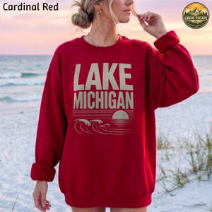 K&ouml;nnte beinhalten: Ein lang&auml;rmeliges Sweatshirt in Kardinalrot mit dem Text LAKE MICHIGAN in gro&szlig;en beigefarbenen Buchstaben. Unter dem Text befindet sich eine Grafik einer untergehenden Sonne &uuml;ber stilisierten Wellen. Das Sweatshirt hat einen Rundhalsausschnitt.