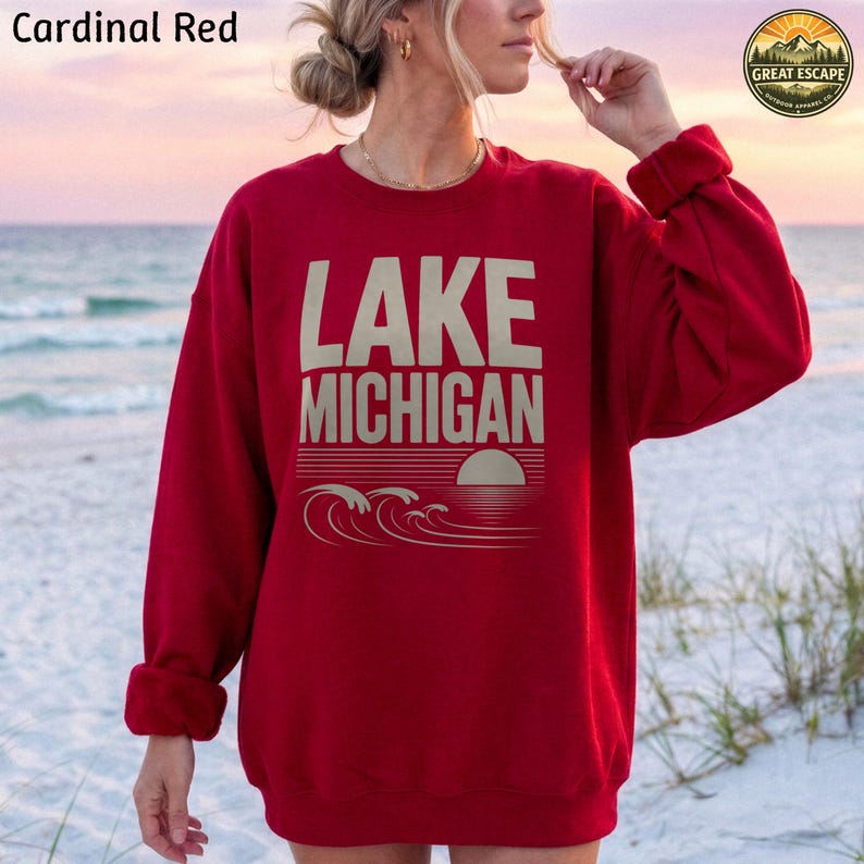 K&ouml;nnte beinhalten: Ein lang&auml;rmeliges Sweatshirt in Kardinalrot mit dem Text LAKE MICHIGAN in gro&szlig;en beigefarbenen Buchstaben. Unter dem Text befindet sich eine Grafik einer untergehenden Sonne &uuml;ber stilisierten Wellen. Das Sweatshirt hat einen Rundhalsausschnitt.