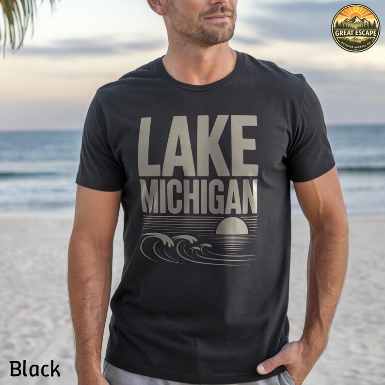 K&ouml;nnte beinhalten: Ein schwarzes kurz&auml;rmeliges T-Shirt mit dem Text LAKE MICHIGAN in gro&szlig;en, hellen Buchstaben &uuml;ber einer Grafik aus stilisierten Wellen und einer Sonne. Ein kreisf&ouml;rmiges Logo in der oberen rechten Ecke enth&auml;lt den Text GREAT ESCAPE und ein Bergmotiv.