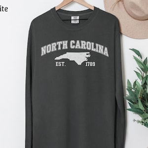 May include: Graphite long-sleeve sweatshirt with "NORTH CAROLINA" arched across the chest in white, with a white outline of the state and "EST. 1789" below. The shirt is hanging on a wooden hanger.