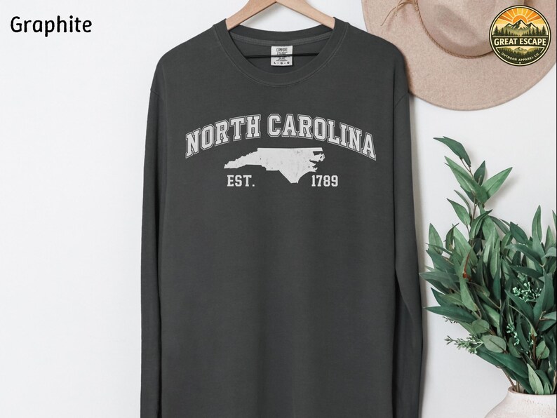May include: Graphite long-sleeve sweatshirt with "NORTH CAROLINA" arched across the chest in white, with a white outline of the state and "EST. 1789" below. The shirt is hanging on a wooden hanger.