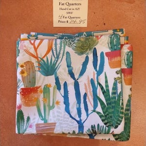 Könnte beinhalten: Gefaltete Stoff-Fat Quarters mit einem südwestlichen Kaktusdruck in Grün-, Blau-, Orange- und Gelbtönen auf weißem Hintergrund. Der Stoff ist mit "Southwestern Fat Quarters" und "Hand Cut in AZ!" beschriftet.