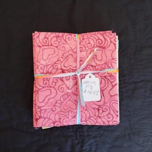 May include: A bundle of pink batik fabric squares, patterned with swirling heart shapes in a darker pink. The fabric is tied with ribbons. A tag with handwritten text is attached.