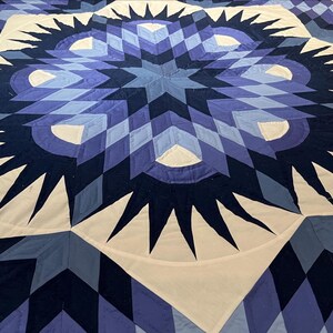May include: A large, handmade quilt with a striking geometric design. The quilt features a central starburst pattern in shades of blue, from light to dark, with white accents. The design is composed of diamonds, triangles, and curved shapes.