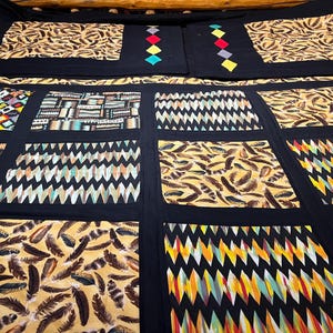 May include: A patchwork quilt with a black background and various patterned squares. Some squares feature feather designs, while others have geometric patterns in colors like yellow, orange, and turquoise. The quilt also has decorative diamond shapes.