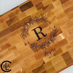 May include: Wooden cutting board with a mosaic pattern. The board features a black floral wreath design around the letter "R" and the name "Rogers" with "EST. 2011" below. The board is made of light and dark wood.