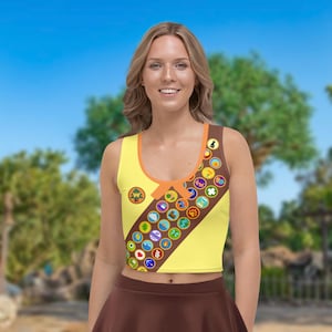 May include: Yellow sleeveless crop top with a brown sash and colourful patches. The sash has the text "Wilderness Explorer" and a tree logo.