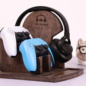 Peut inclure: Station de jeu en bois marron foncé avec un support pour deux manettes de jeu, un casque et un petit plateau. Le support porte le nom "RICHARD". Une manette blanche et une bleue sont dans le support, et un casque noir est accroché sur le côté.