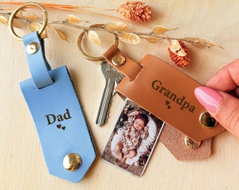 May include: Two leather keyrings, one light blue and one tan, with gold hardware. The blue keyring says "Dad" and the tan keyring says "Grandpa". A photo of a baby is attached to the tan keyring. Dried flowers are in the background.