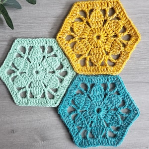 May include: Three handmade crochet hexagon coasters in shades of yellow, teal, and mint green. Each coaster features a floral design in the center and an openwork border. The coasters are arranged on a light gray wooden surface.