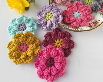 May include: A collection of seven crocheted flower embellishments in various colors, including pink, purple, blue, yellow, and red. The flowers are arranged on a white surface.