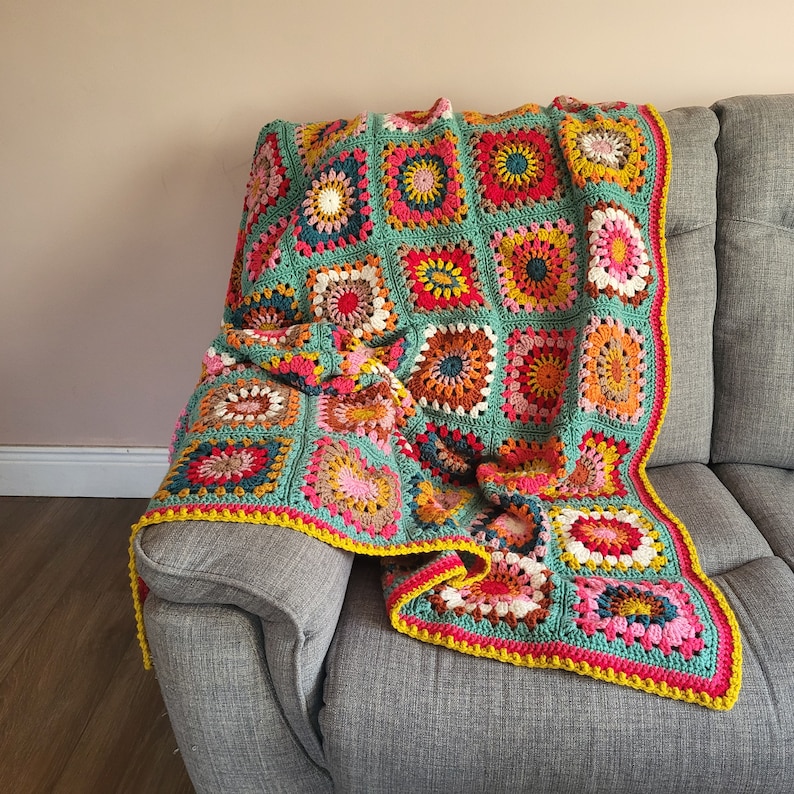 May include: A vibrant, hand-crocheted blanket in a patchwork design, draped over a grey sofa. The blanket features square motifs in teal, pink, yellow, orange, and brown. The edge is trimmed with a bright yellow border, adding a pop of colour.