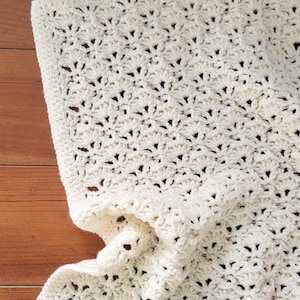 May include: Cream-colored, handmade crochet blanket. The blanket features a detailed, openwork pattern with a scalloped edge. The soft, textured blanket is folded, revealing the intricate stitchwork and cozy design. Perfect for a baby or as a decorative throw.