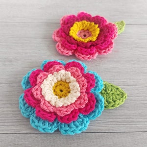 May include: Two crocheted flowers with green leaves. The larger flower has layers of blue, pink, and white crocheted petals with a yellow center. The smaller flower has pink petals with a yellow center.