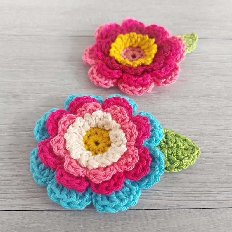 May include: Two crocheted flowers with green leaves. The larger flower has layers of blue, pink, and white crocheted petals with a yellow center. The smaller flower has pink petals with a yellow center.