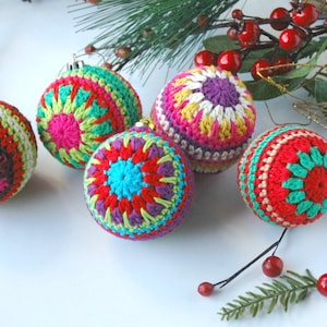 May include: A collection of colorful, crocheted Christmas ornaments. The ornaments feature vibrant patterns in shades of red, pink, green, blue, and purple. They are displayed with sprigs of greenery and red berries.