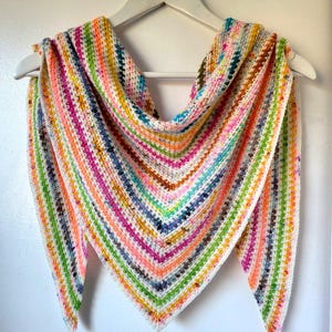 May include: A colorful, handmade crochet shawl with a striped pattern. The shawl features a variety of bright colors including pink, green, orange, and blue. It is displayed on a white hanger against a white background. The shawl is triangular in shape.