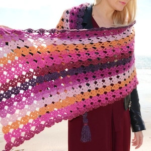 May include: A colorful crocheted shawl with a gradient of pink, purple, and orange hues. The shawl has a delicate lace pattern and a tassel at the end.