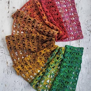 May include: Overhead shot of five crocheted scarves in various colors. The scarves are arranged on a white, distressed wooden surface. Colors include shades of red, orange, brown, yellow, and green. The scarves have an open, lacy design.