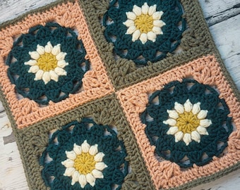 May include: A square crochet textile composed of four smaller squares joined together. Each square features a central yellow flower with white petals, surrounded by dark teal yarn, framed by either olive green or light peach borders.