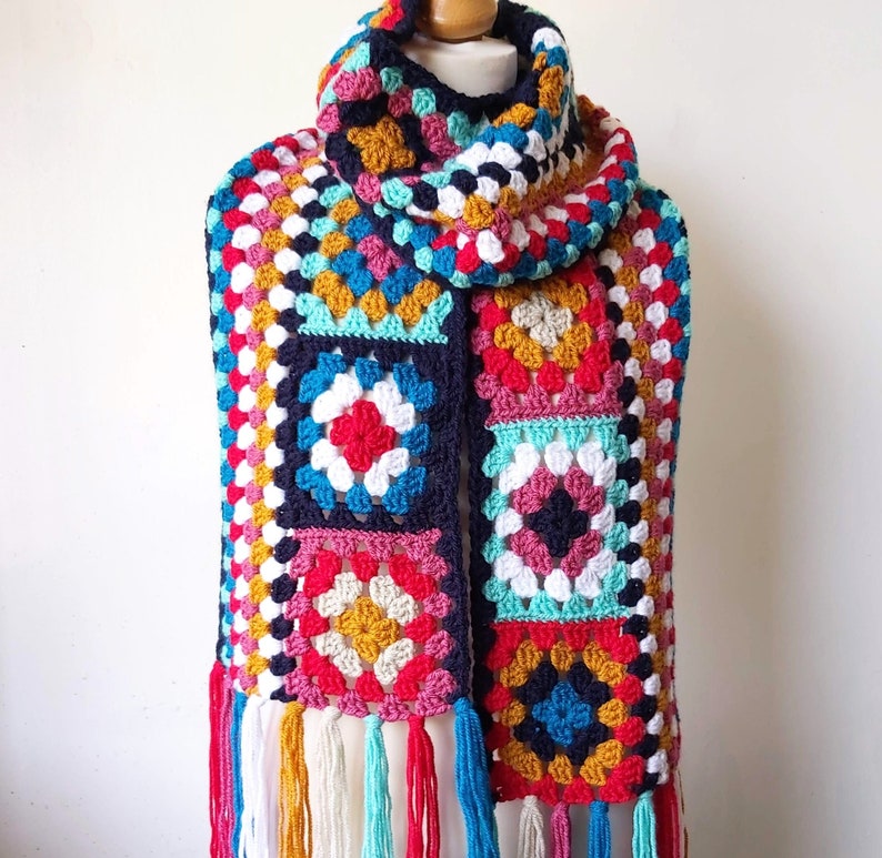 May include: A colorful crocheted scarf with a fringe. The scarf is made of squares in various colors, including red, blue, green, yellow, and white. The scarf is tied around a mannequin's neck.