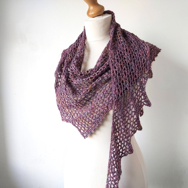 Puede incluir: Un chal de ganchillo morado y marr&oacute;n con un dise&ntilde;o de encaje. El chal est&aacute; drapeado sobre un maniqu&iacute; blanco.