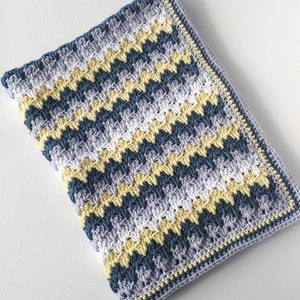 Könnte beinhalten: Eine gehäkelte Babydecke mit einem Chevron-Muster in Blautönen, Gelb und Weiß. Die Decke hat einen gehäkelten Rand in Gelb und Weiß.