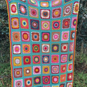 May include: A vibrant, hand-crocheted blanket. It has a turquoise base with many square patterns in pink, yellow, orange, and brown. The blanket is edged with a yellow border and a red trim.