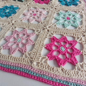 May include: A crocheted blanket with a beige background and colorful flower designs. The blanket has a pink, blue, and green border.