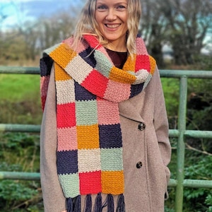 May include: A crochet scarf featuring a multi-colored checkerboard pattern in shades of mustard yellow, navy blue, light pink, red, and sage green. The scarf is finished with long, dark navy blue fringe tassels at both ends of the rectangular knit fabric.