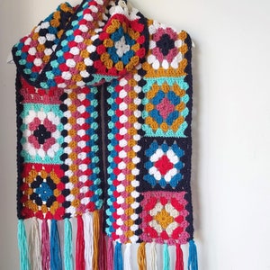 May include: A colorful crocheted scarf with a geometric pattern in shades of red, blue, green, yellow, and white. The scarf has a fringe on the bottom and is made of a soft, textured yarn.